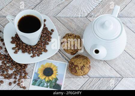 Two chocolate drops cookies surrounded by cup of coffee, teapot and a picture of a sunflower. On a wooden table (top view). Stock Photo