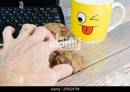 Male hand picking up a cookie with drops of chocolates. In the background, a yellow mug and a laptop. Pause for e-learning. Stock Photo