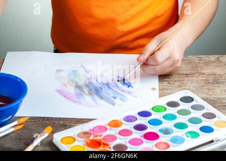 A caucasian boy is drawing an abstract picture on a white paper using a set of watercolor. He is left handed and holding brush. Artistic development, Stock Photo
