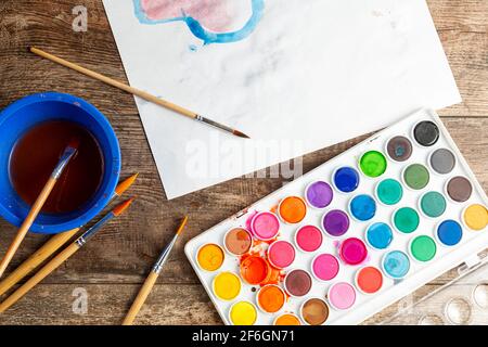 Close up top view image of a wooden desk with various sizes of brushes a cup of water, a messy watercolor palette and a blank paper for drawing. Art c Stock Photo