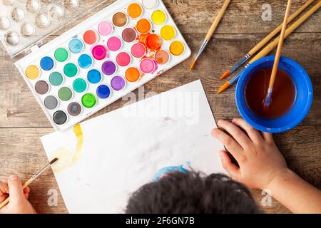 A caucasian boy is drawing an abstract picture on a white paper using a set of watercolor. He is left handed and holding brush. Artistic development, Stock Photo