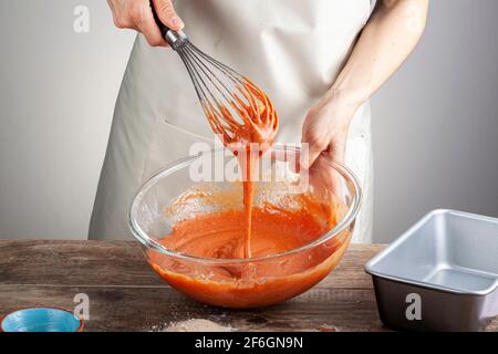 A woman is mixing a blood orange cake mix in a glass bowl using a balloon whisk. Concept for cooking, homemade recipe, food coloring, additives. Stock Photo