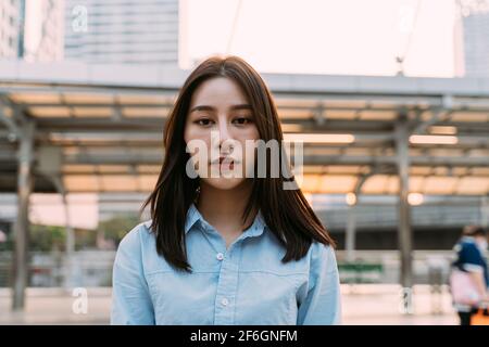 Portrait of thoughtful young Asian businesswoman standing looking at camera feeling depressed and unhappy standing in city street Stock Photo