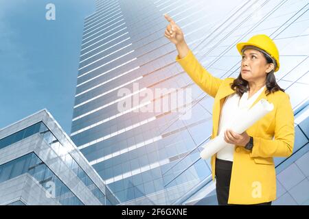 Asian engineer woman with blueprint standing at building background with copy space, engineering construction and architecture concept. Stock Photo