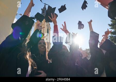 Congratulations Low angle view of happy group of six young cheerful graduates in black gowns, throwing up their head wear in the air and celebrating, Stock Photo