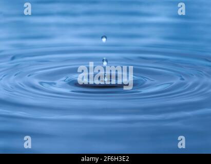Water droplet dripping into blue pool creating ripples and abstract shape of water. Darkest area in center is in camera focus. Image contains some gra Stock Photo