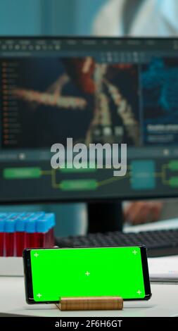 Microbiologist bringing blood samples in modern lab putting them near smartphone working with green chroma key screen placed on desk. Team of biotechnology scientists developing drugs in background Stock Photo