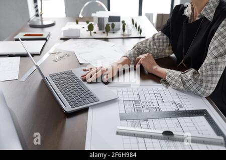 Close-up of female architect sitting at her workplace with blueprints and using laptop at work at office Stock Photo