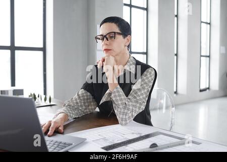 Mature businesswoman in eyeglasses sitting at the table and using laptop computer in her online work Stock Photo