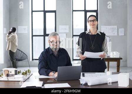 Portrait of successful architects looking at camera while working with blueprints and computer at the table at office Stock Photo