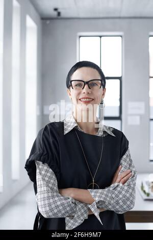 Portrait of mature businesswoman in eyeglasses smiling at camera while standing with arms crossed at office Stock Photo
