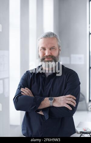 Portrait of mature bearded businessman smiling at camera while standing with his arms crossed at office Stock Photo