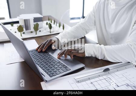 Close-up of African architect sitting at the table with blueprints and typing on laptop computer Stock Photo