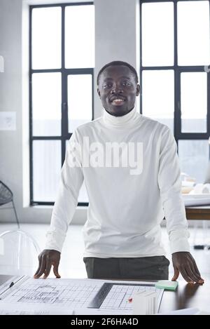 Portrait of African young architect smiling at camera while standing at his workplace with blueprints at office Stock Photo