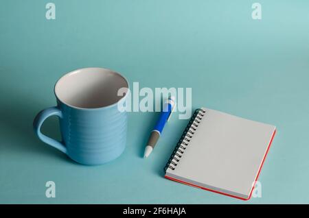 Blue striped mug, pen and open notebook on a blue background. Empty new ceramic mug, blue plastic pen, and blank spring notepad page. Close-up. Select Stock Photo