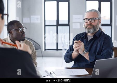 Mature bearded businessman sitting at meeting and talking to his colleagues during work at office Stock Photo