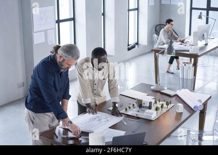 Two architects standing near the table with model of buildings and blueprints and developing new project in team Stock Photo