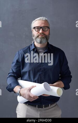 Portrait of mature bearded architect holding blueprint and looking at camera while standing against the black background Stock Photo