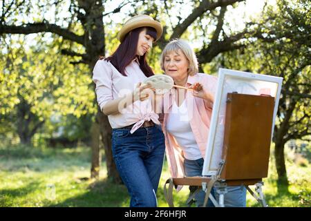 Close up outdoor portrait of senior 60-aged woman and her young daughter, painting together using easel on sunny day in the garden. Elderly mother and young daughter choosing a paint on a palette. Stock Photo