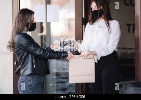 Young woman standing outdoors in mask and paying bill through smartphone using NFC technology in a cafe. Female customer paying using contactless Stock Photo