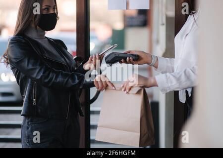 Young woman standing outdoors in mask and paying bill through smartphone using NFC technology in a cafe. Female customer paying using contactless Stock Photo