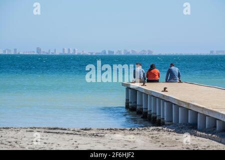 Three people sitting on the end of a jetty in the Mar Menor at Los Alcazares, Murcia, Spain Stock Photo