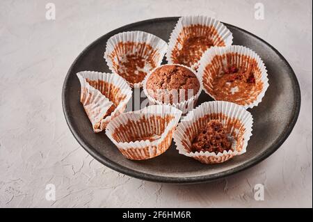 Homemade chocolate cupcake in baking paper form, next to empty wrappers on dark plate on wooden background, selective focus Stock Photo