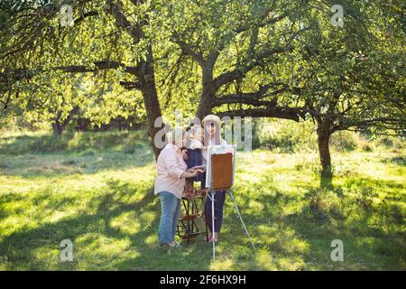 Beautiful summer spring shot of happy three generations family, little kid girl, young mother and mature grandmother, spending time outdoors and painting picture on easel. Summer family leisure. Stock Photo