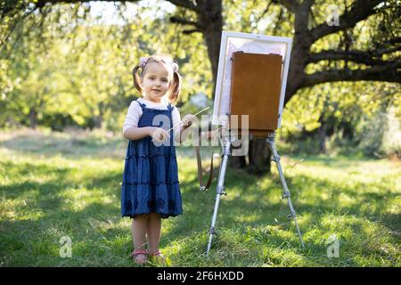 Little smiling girl in jeans dress, painting at green summer park outdoor. Talented small girl artist, holding brush in hands, posing to camera near the easel at sunny spring garden. Stock Photo
