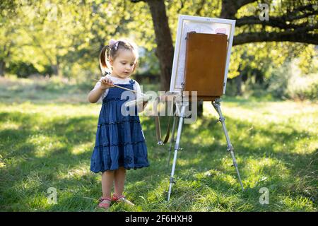 Painting on easel outdoors in the nature. Little cute talented excited kid girl artist, painting a picture on canvas, holding palette and brush and choosing a paint color. Stock Photo