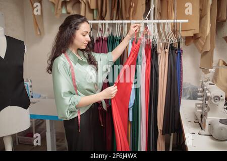 Female fashion designer working on new womenswear collection for clients in cozy workshop studio, dressmaker, tailor or needlewoman standing near