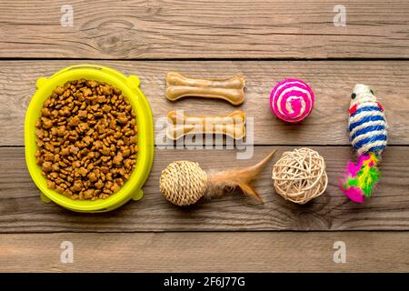 Dry food in bowl, spatula, collar, mouse, balls, bones on wooden background Top view Flat lay Toys, accessories and food for your beloved pet concept. Stock Photo