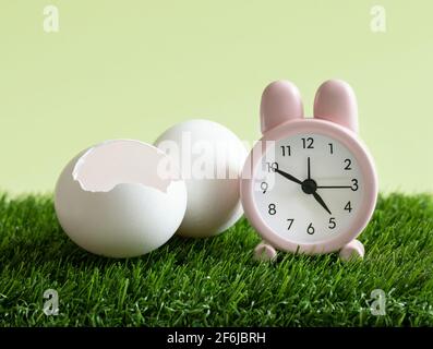 Easter time concept. Little pink rabbit alarm clock and white eggs on the green grass background. Countdown to the holiday. The hatched egg. Stock Photo