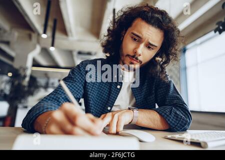 Handsome curly guy is making notes at the working table in open space office Stock Photo