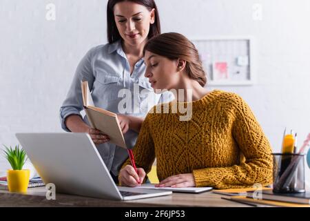 teenage girl writing while doing homework near laptop and mother with book Stock Photo