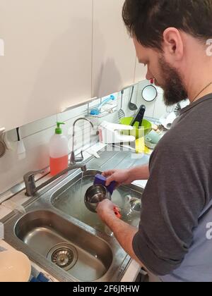 a bearded tired and nervous man washes dishes in the kitchen sink after dinner Stock Photo