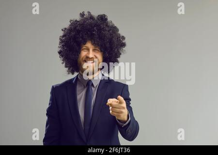 Toothy smiling businessman in curly toupee pointing to camera studio portrait Stock Photo