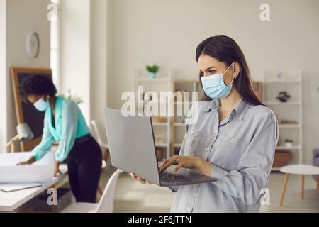 Woman employee wearing facial mask using laptop stand keep distance in office Stock Photo