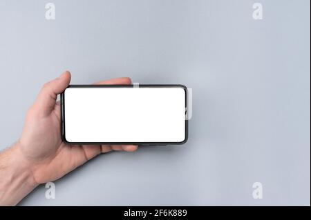 Mockup cell phone. male hand holding phone horizontally with blank white screen. grey table background. Mockup phone horizontal. human left hand holdi Stock Photo