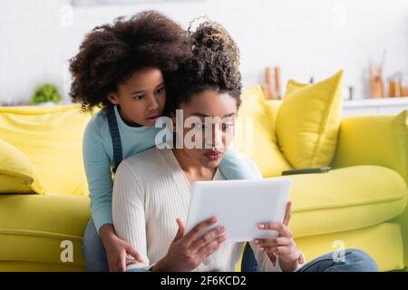 surprised african american girl with mom looking at digital tablet
