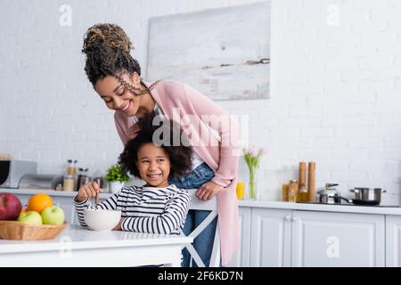 happy african american woman standing near smiling daughter having breakfast in kitchen Stock Photo