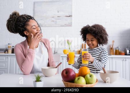 smiling african american woman covering mouth while daughter holding orange juice in kitchen, blurred foreground Stock Photo