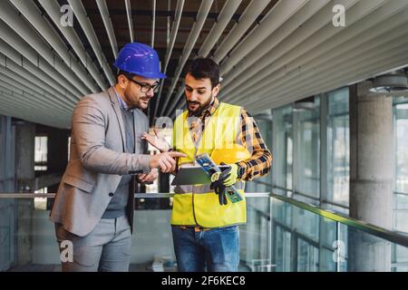 Architect and construction worker standing in building in construction process and looking at blueprints on tablet. Stock Photo