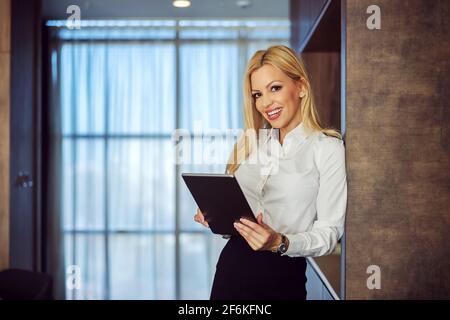 Corporate photography. Pretty blonde smiling woman, well dressed with her arm against the wall, holds a digital tablet in her hands and smiles at the Stock Photo