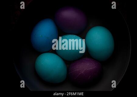 Blue, turquoise, and purple Easter eggs in a bowl with strong shadows and dark background. Stock Photo