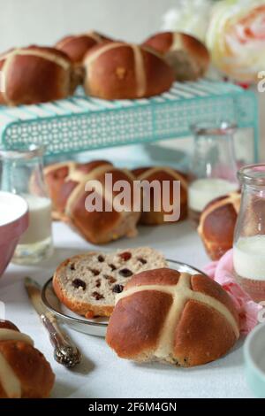 Lots of freshly baked homemade Hot Cross Buns on a table with small bottles of milk and one bun split open ready to be spread with butter Stock Photo