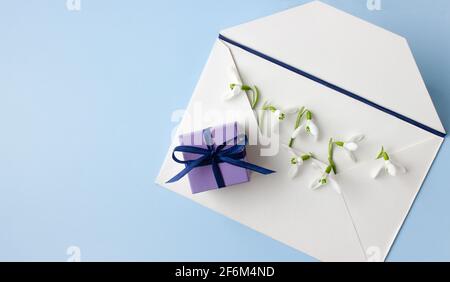 Beautiful snowdrops in white envelope and purple gift box on light blue background, closeup, space for text, top view. Stock Photo
