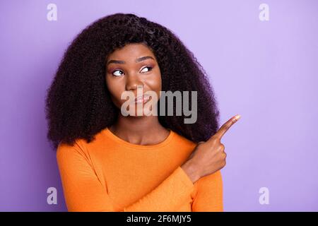 Photo of sweet unsure dark skin lady wear orange shirt looking ponting empty space isolated violet color background Stock Photo