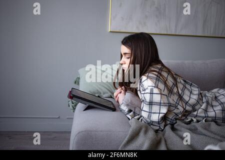 Caucasian young woman looks at the tablet lying on couch in modern home and using wireless internet. Female learn online. Stock Photo
