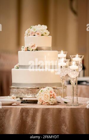 Wedding ceremony three tiered cake made with white frosting and grey trim decorated with white and pink roses on a table covered with silk tablecloth Stock Photo